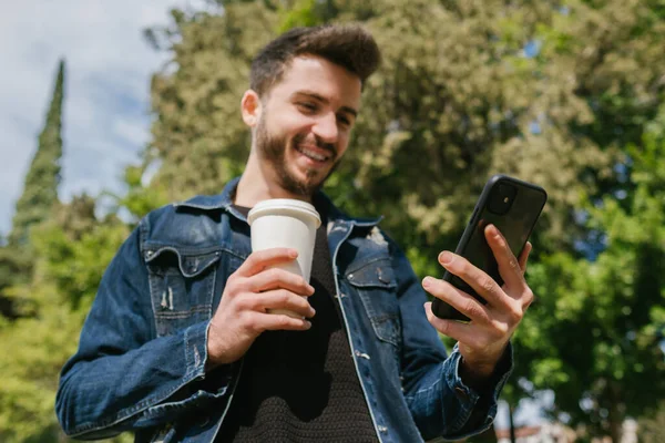 Açık havada elinde kağıt kahve fincanı tutarken cep telefonu ekranına bakan kot pantolon giyen mutlu genç adam. Sosyal medyada kaydırma, video izleme veya arkadaşlara mesaj gönderme.
