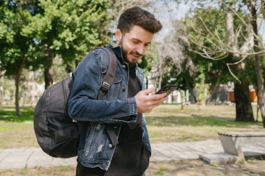 Sırt çantası olan yakışıklı bir öğrenci ve dışarıda cep telefonu kullanıyor. Kaydırma, mesaj okuma veya video izleme.