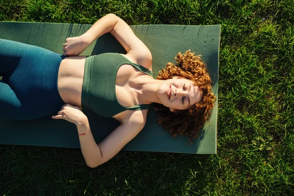 Yeşil spor sutyen ve mavi yoga pantolonu giyen güzel, genç ve sportif bir kadın manzarası. Yoga minderinde yatarken gözleri kapalı ve gülümsüyor..