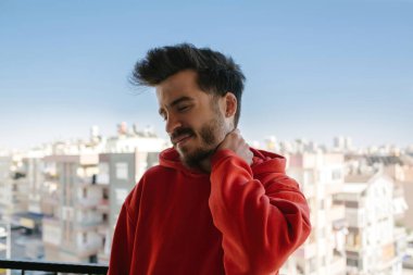 Bitkin hissediyorum. Balkonda boynu ağrıyan kırmızı kapüşonlu mutsuz bir adam. Hüsrana uğramış genç yakışıklı adam metal çitlere yaslanırken bitkin görünüyor. Sağlık ve sorun kavramı.