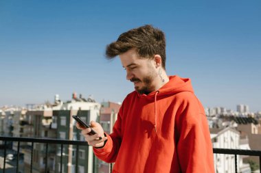 Telefonu kullanarak kırmızı kapüşonlu yakışıklı genç adam. Adam gülümseyerek akıllı telefon ekranı arıyor. Arkadaşla mesajlaşma, video izleme veya sosyal medyada kaydırma.