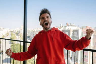 Genç yakışıklı adam, gündelik kırmızı kapüşonlu çok mutlu ve heyecanlı. Kazanan bir jest yapıyor. Kolları havada, gülümsüyor ve başarı için bağırıyor..