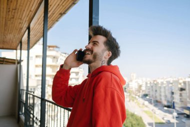 Telefon görüşmesi, kırmızı tişörtlü genç adam balkonda telefonla konuşuyor, gülümseyen yakışıklı yetişkin.