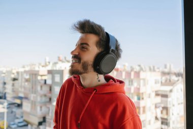 Kulaklık, kulaklıklı kırmızı kazaklı adam balkonda müzik dinliyor, müzik dinliyor ve anın tadını çıkarıyor.