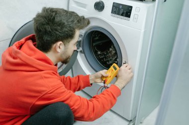 Kırmızı kapüşonlu genç adam evde dijital çoklu sayaçla çamaşır makinesini kontrol ediyor..