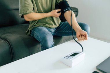 Young man wearing khaki tees and denim pants using modern tonometer at home. Cropped of middle-eastern man measuring his blood pressure, making self checkup.