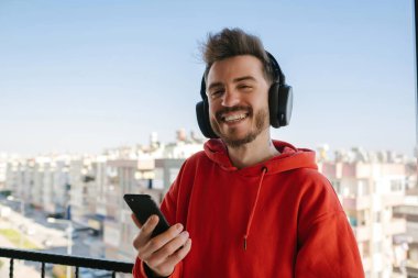 Kırmızı kapüşonlu genç adam elinde akıllı telefon ya da cep telefonu tutarken kameraya gülümsüyor. Cep telefonu kullanılıyor.