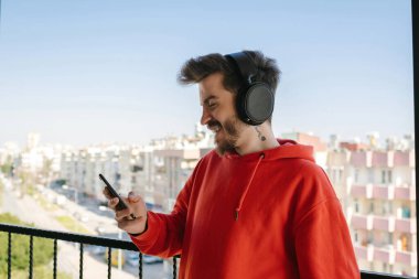 Kırmızı kapüşonlu yakışıklı genç adam kulaklıklı akıllı telefonla video görüşmesi yapıyor. Telefonla video izleyen ya da sosyal medyada kayan bir adam.