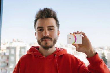 Kırmızı kapüşonlu genç adam kredi kartı ya da banka kartı gösteriyor. Çevrimiçi alışveriş veya dijital bankacılık konsepti. Erkeğe odaklan.