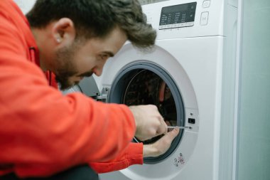 Tamirci, genç yakışıklı adam evindeki bozuk çamaşır makinesini tamir ediyor, tornavidayla vidaları sıkıyor.