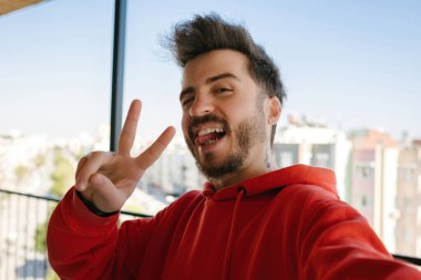 Poz veren, selfie çeken ve iki el hareketi yapan genç adam, kırmızı kazak giymiş mutlu yetişkin erkek, eğlence ve neşe