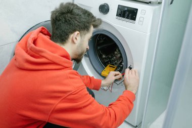 Tamirci, genç yakışıklı adam evindeki bozuk çamaşır makinesini tamir ediyor. Voltmetreyle elektrik olup olmadığını kontrol ediyor.