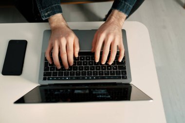 Masadaki dizüstü bilgisayarda, genç adam makale yazıyor, bilgisayarındaki postalarını kontrol ediyor, beyaz bir masada oturuyor, uzaktan çalışıyor ve oturma odasından iş yapıyor.