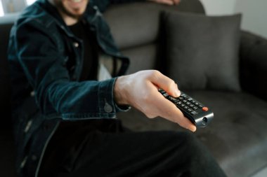 TV remote, young man sitting on sofa in living room changing channels with remote in hand, video streaming services and TV series viewing sites