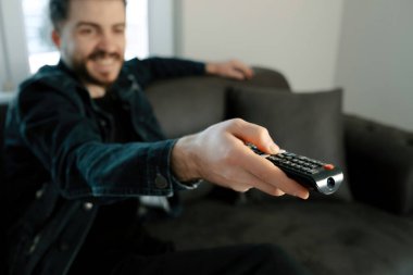 Uzaktan kumandalı kot elbise giyen mutlu genç adam evde TV 'de kanal değiştiriyor. Mutlu genç adam TV 'de ilginç bir program izliyor..
