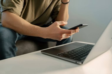 Phone on hand, adult man is holding his phone, laptop is on the table and he is browsing social networking sites, young man is checking his mails