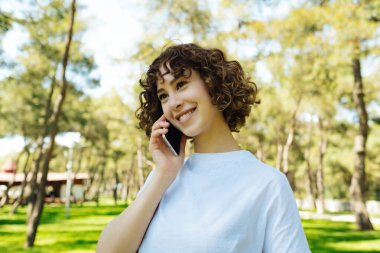 Talking phone, cheerful pretty young redhead woman talking on phone and look straight ahead. Holding smartphone close to ear. Standing alone outside.
