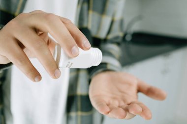 Young man wearing lumberjack shirt taking pill. Hand spilling pills for the pain of a bottle in the kitchen. Treatment, medicine, medication, pills, vitamins, supplements concept.