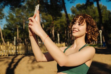 Spor eğitmeni Koç Hanım 'ın şehir parkında, açık havada video selfie kaydeden akıllı cihazının fotoğrafı. Genç kızıl saçlı kadın telefonuyla selfie çekiyor. Açık hava sporları ve sağlıklı yaşam kavramları.