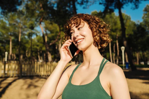 Şehir parkında, açık havada duran neşeli kızıl saçlı bir kadının görüntüsü. Yeşil spor sutyen takmış cep telefonuyla konuşuyor ve gülümsüyor..
