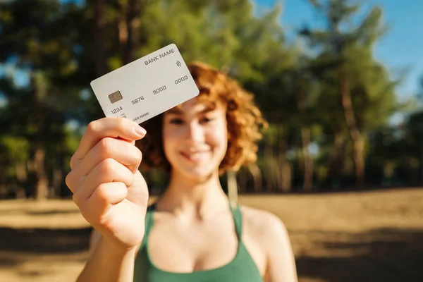 Şehir parkında, açık havada dururken kredi kartı gösterirken gülümseyen yeşil spor sutyen giyen neşeli genç bir kadın. Çevrimiçi alışveriş, bankacılık, banka kartı. Kredi kartına seçici odaklanma.