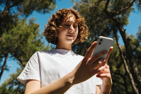 若い赤毛の女性が携帯電話で何かを使用して見ています 白いTシャツを着た若い笑顔の女性の選択的な焦点 緑の都市公園 — ストック写真