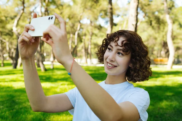 緑の都市公園 屋外で彼女の携帯電話で水平写真を撮る白いティーを身に着けている赤毛の女性 風景写真 屋外のライフスタイルの概念 — ストック写真