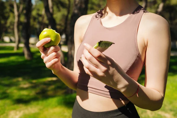 Elma yiyen ve egzersiz yaparken protein çubuğu tutan güzel sporcu kadın. Fitness kızı için sağlıklı bir atıştırmalık. Fitness güzel kadın dışarıda enerji abur cuburu yiyor..