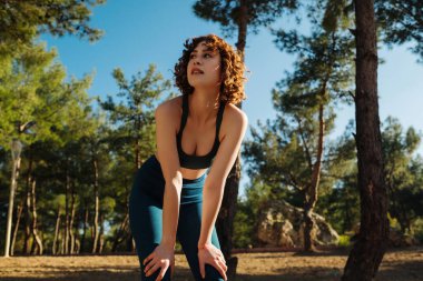 Şehir parkında spor giysiler giymiş genç kızıl saçlı bir kadın, dışarıda duruyor, eğiliyor ve koşu seansından sonra soluklanıyor. Açık hava spor kavramları.
