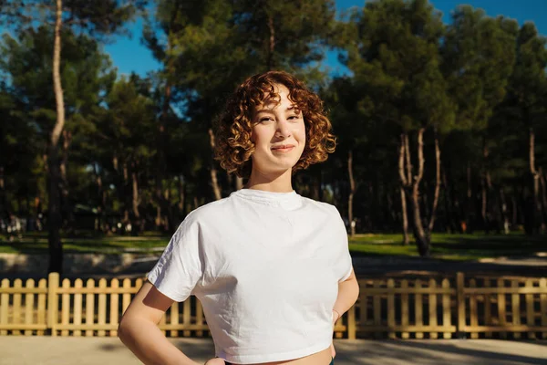 Beyaz tişört giyen, kameraya gülümseyen, şehir parkında, açık havada duran tatlı kızıl saçlı kadın..