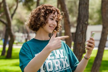 Yeşil tişört giyen, şehir parkında dikilen, elinde telefon tutan ve telefonda gülen yüzlü, gülümseyen genç, kızıl saçlı kadının yan görüntüsü. Cep telefonu ekranına bakıyor..