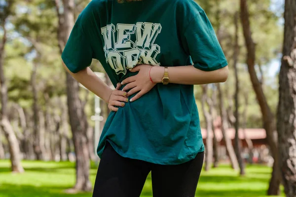 緑のTシャツと黒の足の都市公園に立って着て若いスポーツ女性の作物のビューでは 消化不良 痛みを伴う病気の気分が悪いので 胃の上に手で屋外 アチェの概念 — ストック写真