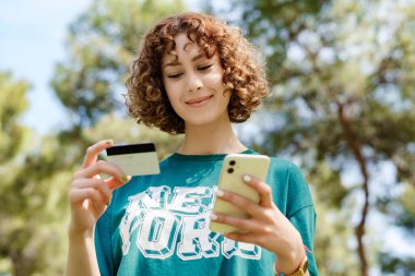 Şehir parkında yeşil tişört giyen mutlu, genç bir kadın elinde cep telefonu kartı olan bir kadın internet üzerinden alışveriş yapıyor ve internet üzerinden ödeme yapıyor. Güvenli mobil bankacılık.