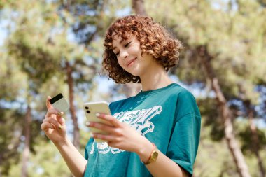 Çevrimiçi alışveriş yapan, şehir parkında duran, elinde cep telefonu kartı tutan yeşil giysili genç, kızıl saçlı bir kadının yan görüntüsü internet üzerinden ödeme sağlıyor. Güvenli mobil bankacılık.