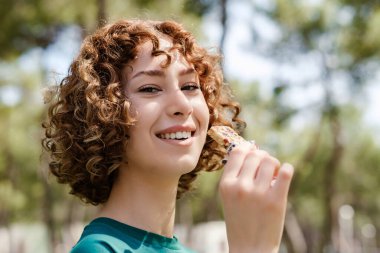 Şehir parkında dikilen yeşil tişörtlü, güzel, mutlu, kızıl saçlı bir kadının protein ya da enerji barını ısırmasını yakından izleyin. Kameraya bakıyorum. Sağlıklı bir yaşam tarzı. Sağlıklı beslenme.