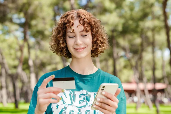 Young happy redhead woman wearing green t-shirt standing on city park, outdoors holding mobile phone credit bank card do online shopping provide internet payment. Safe mobile banking.