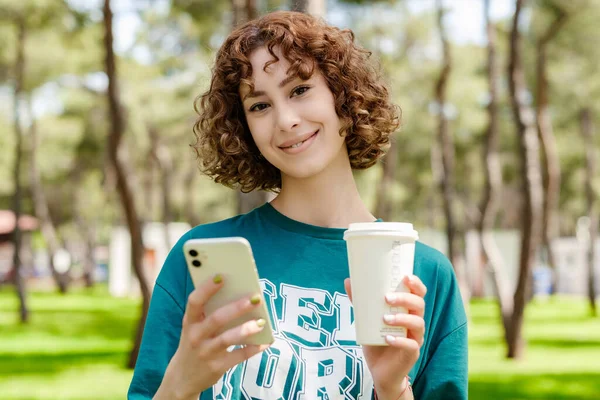 Happy redhead woman wearing casual clothes standing on city park, outdoors holding takeaway coffee mug and using mobile smart phone while looking at the camera. Outdoor sports, healthy life concepts.