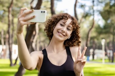 Şehir parkında duran yeşil tişörtlü genç, kızıl saçlı bir kadın akıllı telefonuyla kendi portresini çekiyor. Ekrana bakıyor ve selfie çekiyor..