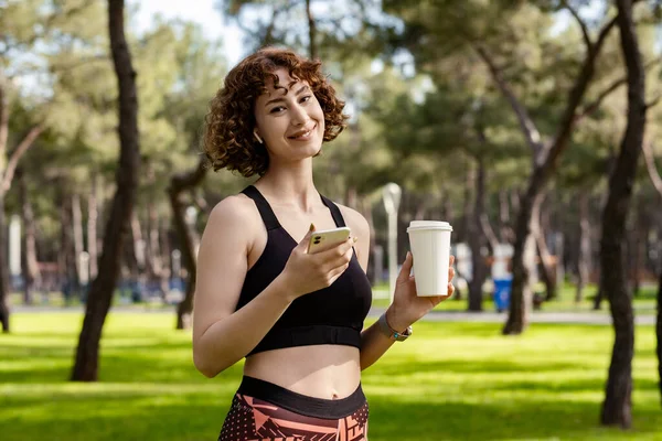 Young woman smiling confident wearing sportive clothes on city park, outdoors holding takeaway coffee mug and using mobile smart phone while looking at the camera. Outdoor sports, healthy life concepts.