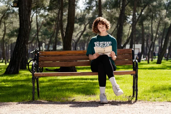 Şehir parkında tişört giyen, açık havada parkta bankta oturan ve kitap okuyan hoş bir kadın. Kişisel gelişim kavramları.