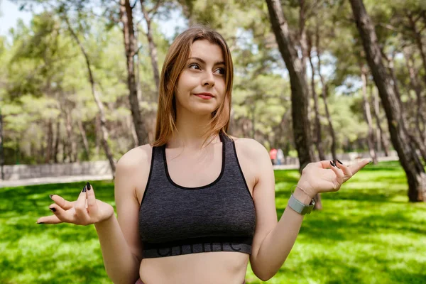Şehir parkında spor sutyen giyen güzel esmer kadın, açık havada habersiz ve kafası karışık, kolları açık, omuzlarını silkiyor, hiçbir fikri yok ve şüpheli bir yüzü var..