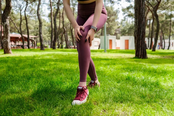 都市公園に立って黒のスポーツブラジャーを身に着けているブルネットのスポーツ女性は 手で屋外で負傷した膝に触れる 足の損傷 健康的なライフスタイルとスポーツの概念 — ストック写真
