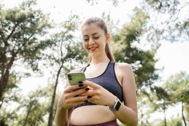 Şehir parkında durmuş telefon ekranına bakıp telefon kullanan siyah spor sutyen giyen hoş bir kadın. Arkadaşlarla mesajlaşma, video izleme veya sosyal medyada kaydırma.
