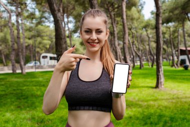 Kentin parkında duran spor sutyenini takan, açık havada cep telefonunu boş beyaz ekranlı gösteren genç bir kadın. Fitness uygulaması. Yeni mobil uygulama öneriliyor, model.