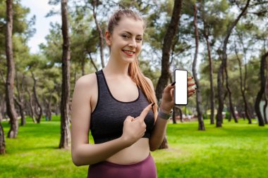 Şehir parkında durmuş siyah spor sutyen giyen, açık havada boş beyaz ekranlı bir cep telefonunu işaret eden hoş bir kadın. Fitness uygulaması. Yeni mobil uygulama öneriliyor, model.