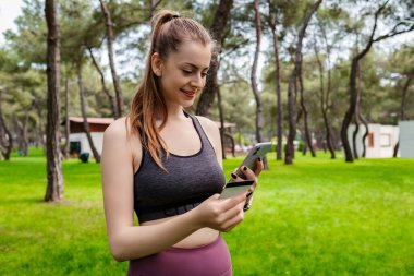 Kentin parkında spor sutyen takan, elinde cep telefonu kredi kartı tutan genç bir kadın internet üzerinden alışveriş yaparak internet üzerinden ödeme yapıyor. Güvenli mobil bankacılık.
