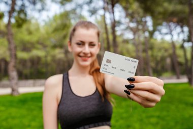Şehir parkında duran spor sutyen giyen mutlu bir kadın, cep telefonu tutarken plastik kredi veya banka kartı gösteriyor. Çevrimiçi alışveriş, bankacılık ve işlem kavramları.