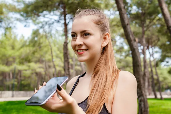 Happy sportive woman wearing sports bra standing on city park, outdoors using her mobile on speaker phone holding the device in front of face. Happy sportive woman talking with friends on phone.