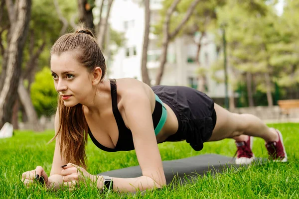 Şehir parkında spor giysiler giyen genç ve güzel bir kadın yoga minderinde tahta egzersizi yapıyor ve dört gözle bekliyor. Sağlıklı yaşam tarzı, egzersiz, eğitim, dayanıklılık konsepti.