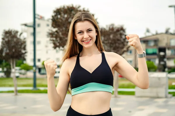 Happy sportive woman wearing sports bra standing on city park, outdoors happy and excited doing winner gesture with arms raised, smiling for success. Feeling triumph, rejoicing goal achievement.